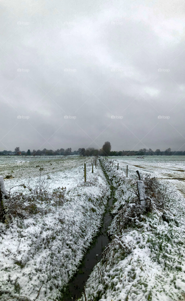 Two farmfields divided by a creek and under a blanket of snow with a grey cloudy snow filled sky above