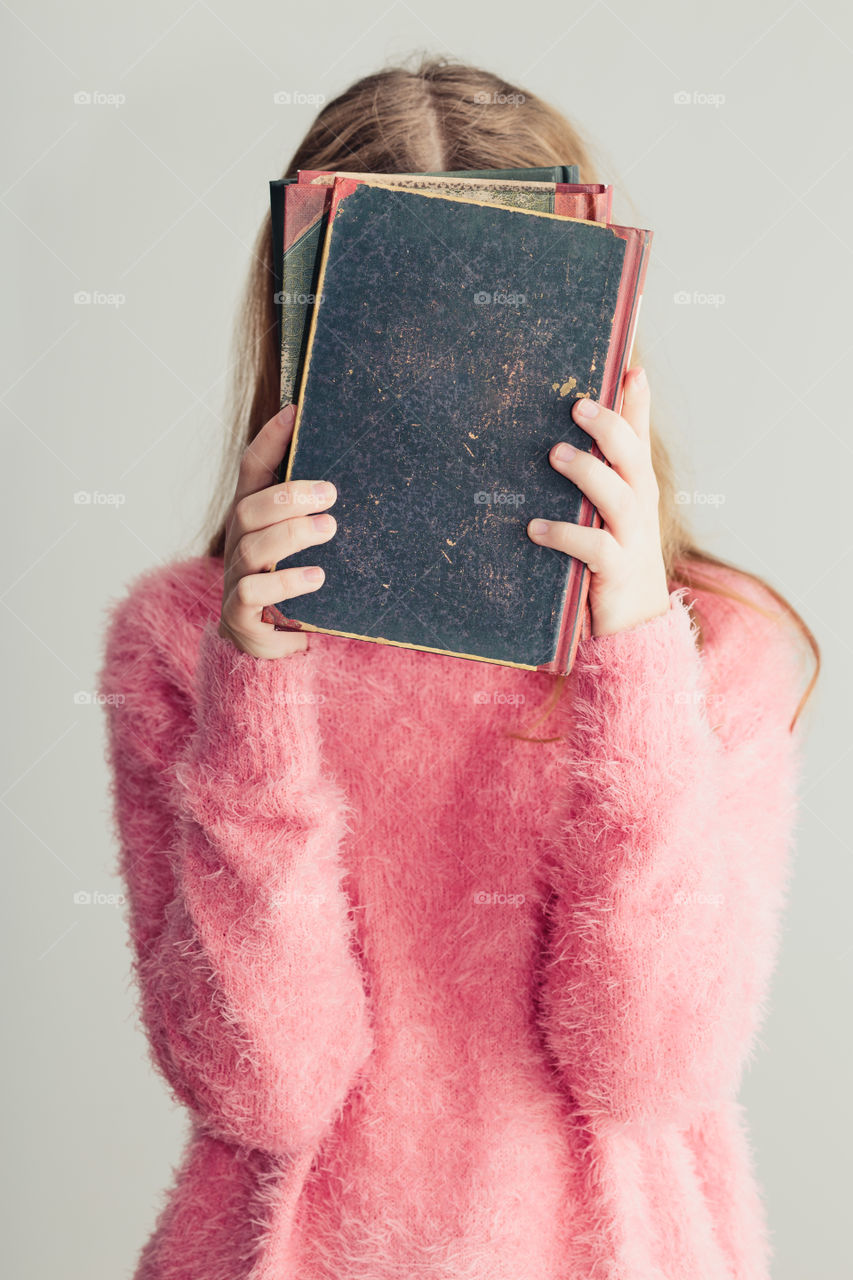 Young girl holding books in front of her face. Teenager girl wearing pink sweater. Vertical photo