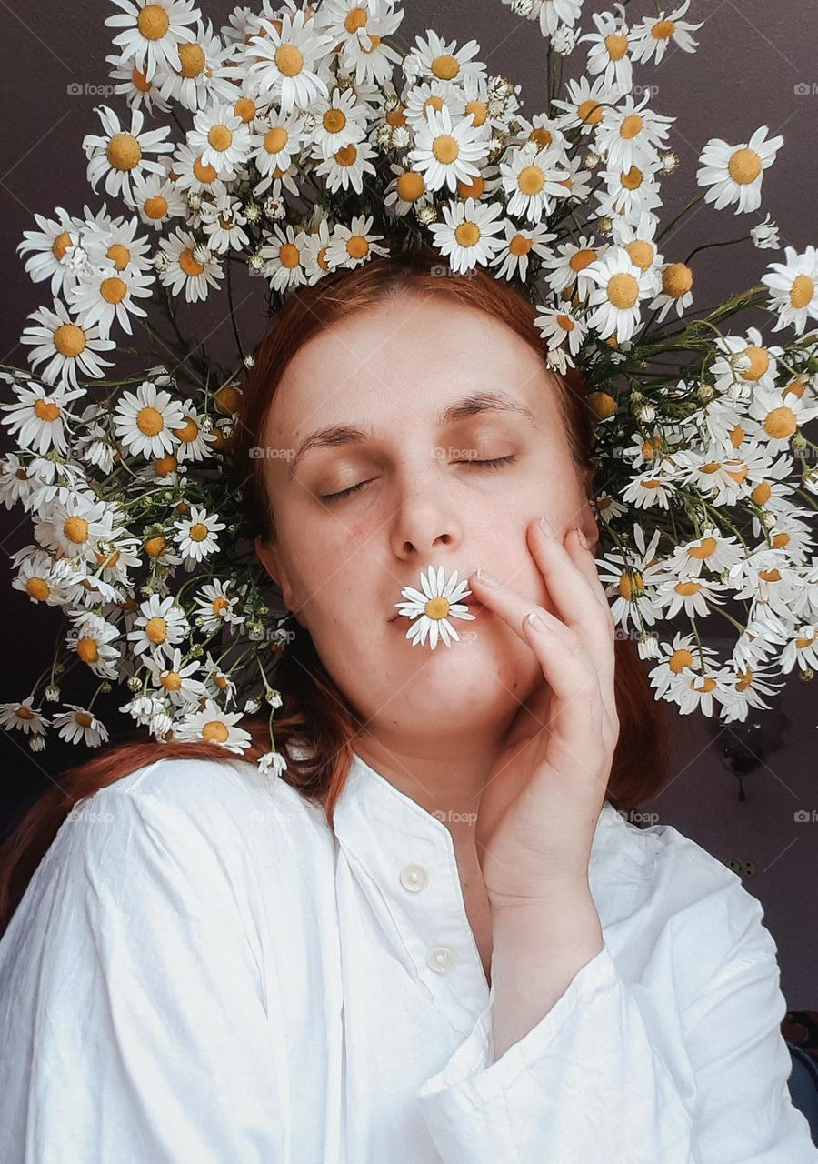 an original portrait of a fair-skinned girl with a wreath of field daisies on her head and one daisy between her lips, symbolizing harmony with nature