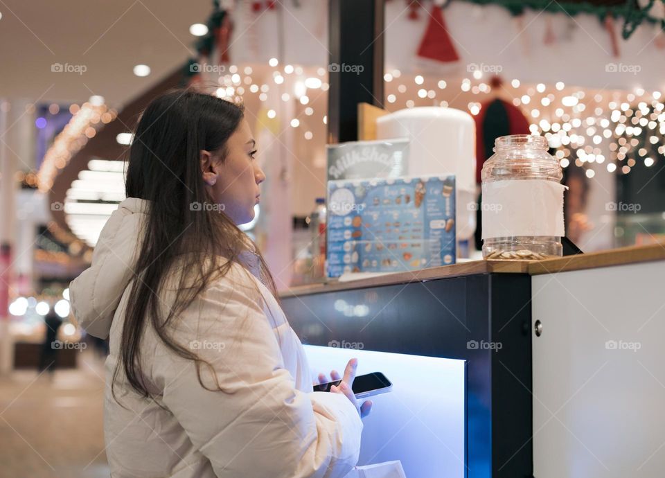 One young beautiful caucasian brunette girl with a phone in her hands stands at the counter with sweets waiting for her dessert in a shopping center in brussels, close-up side view with a blurred background. Holiday shopping offline concept.