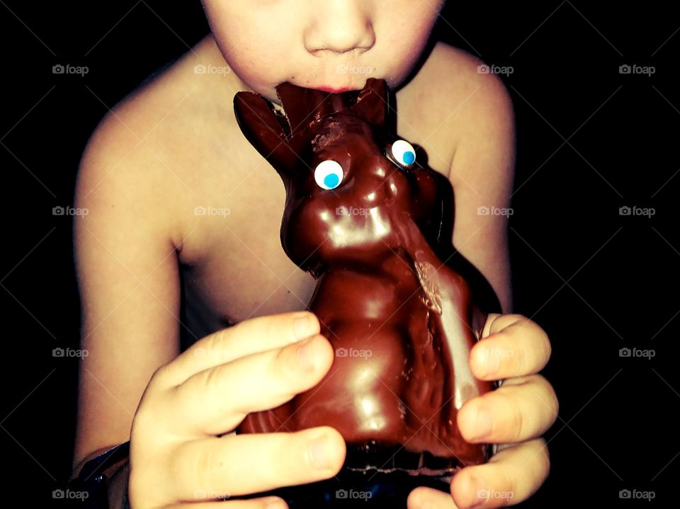 Glowing close up of little boy with his fingers grasping chocolate bunny and he bites into it's ears.