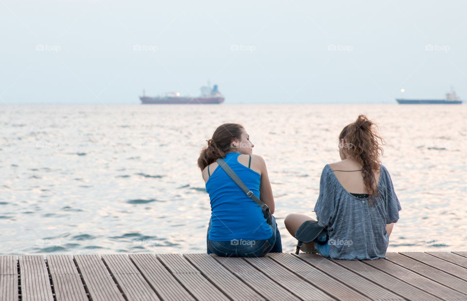 Two Teenagers Girls Friends Talking, Sitting On The Dock And Enjoying The View
