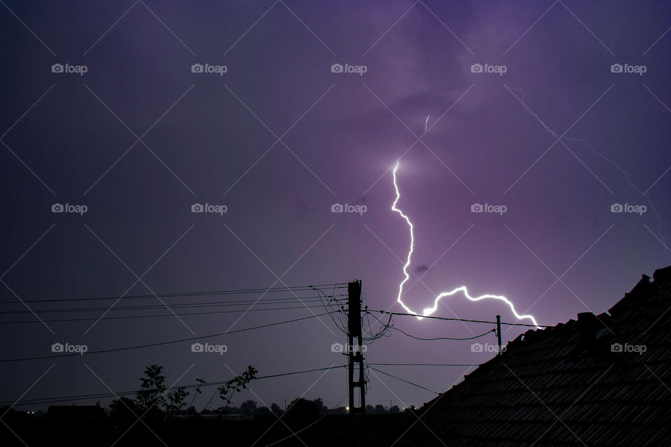 Bright lightning bolt in the sky with a house and powerlines silhouetted against the sky.