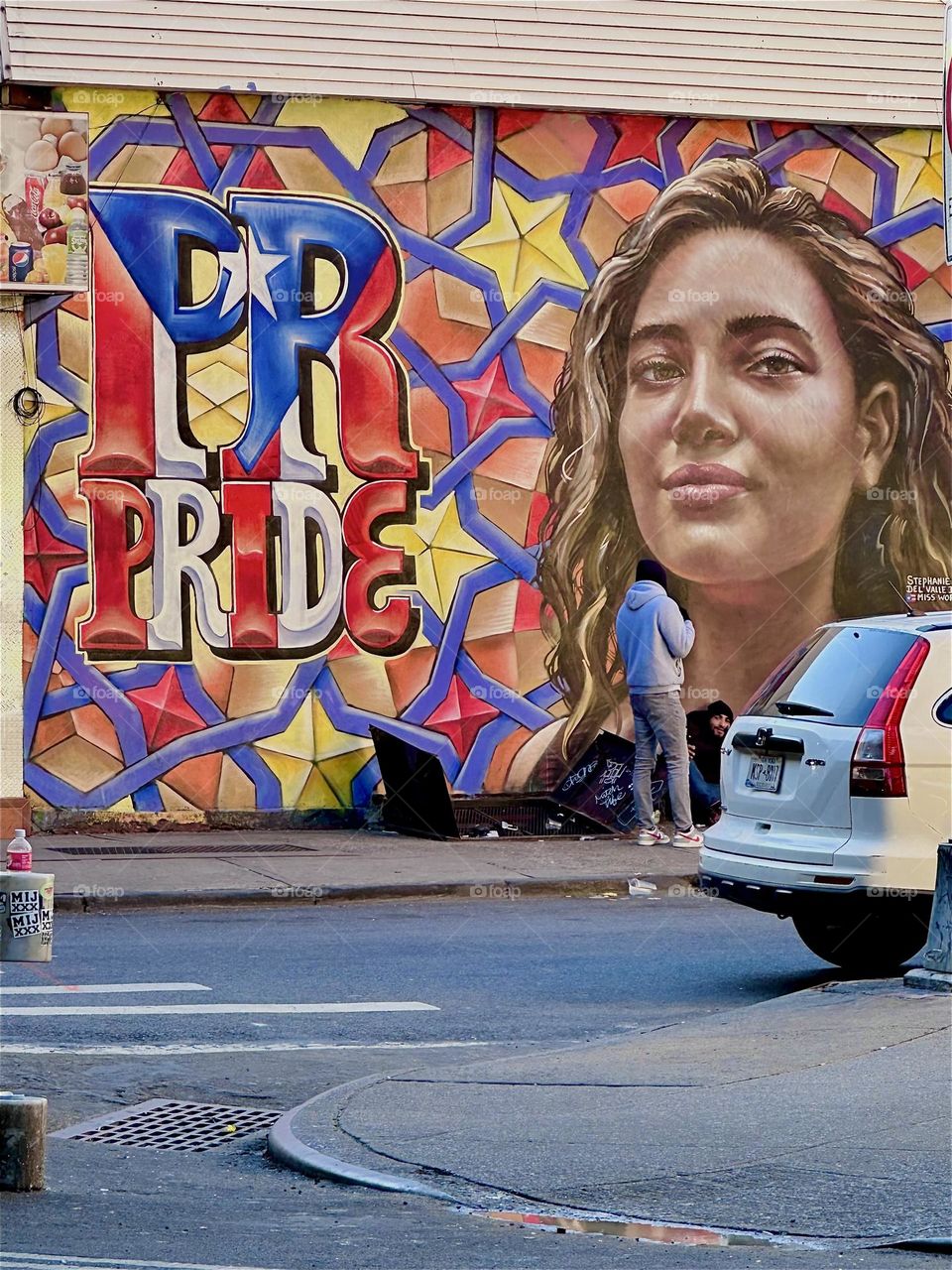 A cool Latina is looking our way from the wall mural next to diamonds and stars and the words “PR” and “PRIDE” on “Jefferson St” and “Broadway” in “Bushwick”, Brooklyn. I am here once again to visit “MG Pharmacy”. 2024. Hypnotic Productions