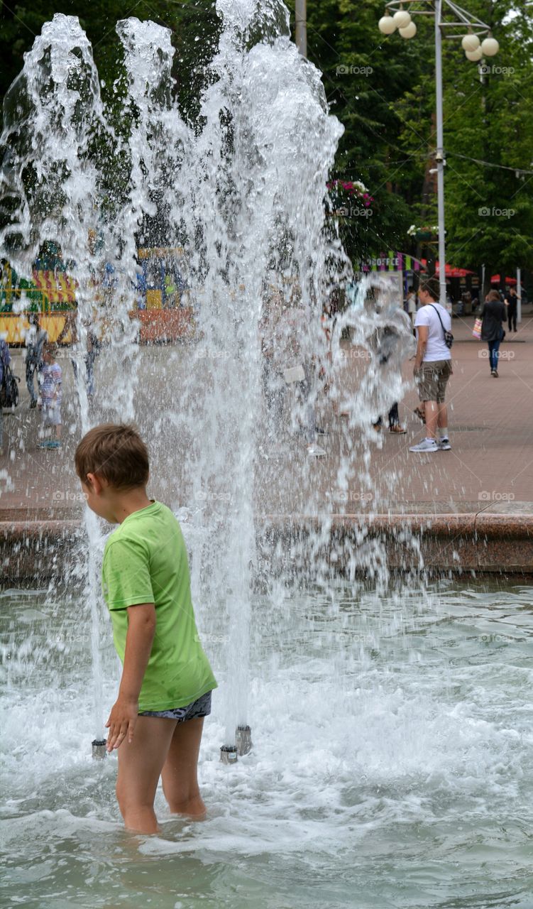 child boy in the water splash fountain, summer heat, city street view