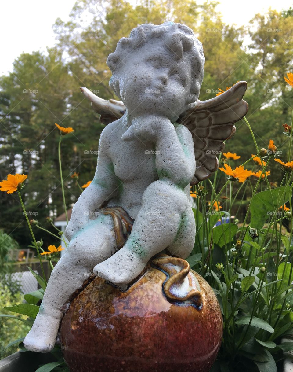 Angel figureine sitting on marble base. Contemplating expression, with flowers, tree greenery & gray sky!👼🏻