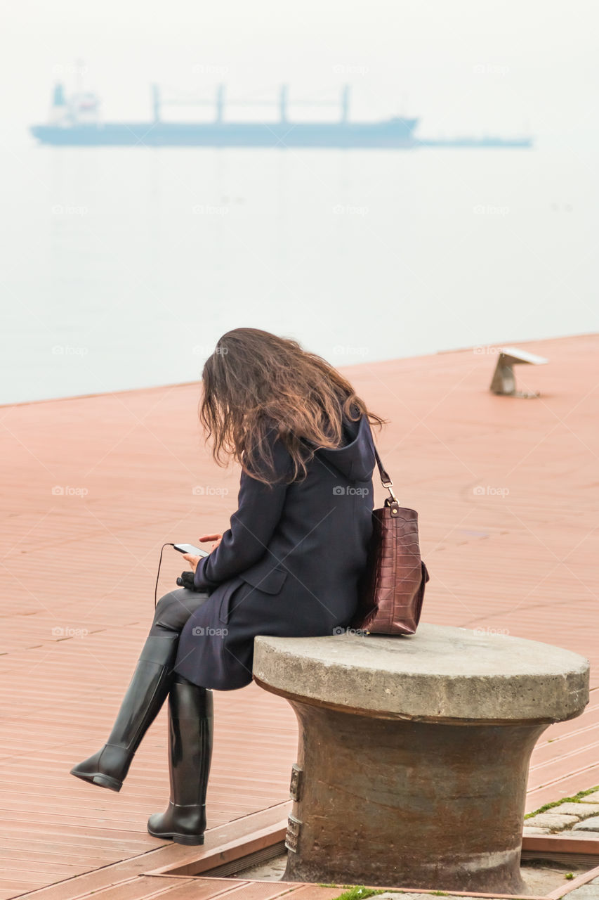 Young Woman Sitting Alone At The Dock Using Her Smartphone And Listening Music
