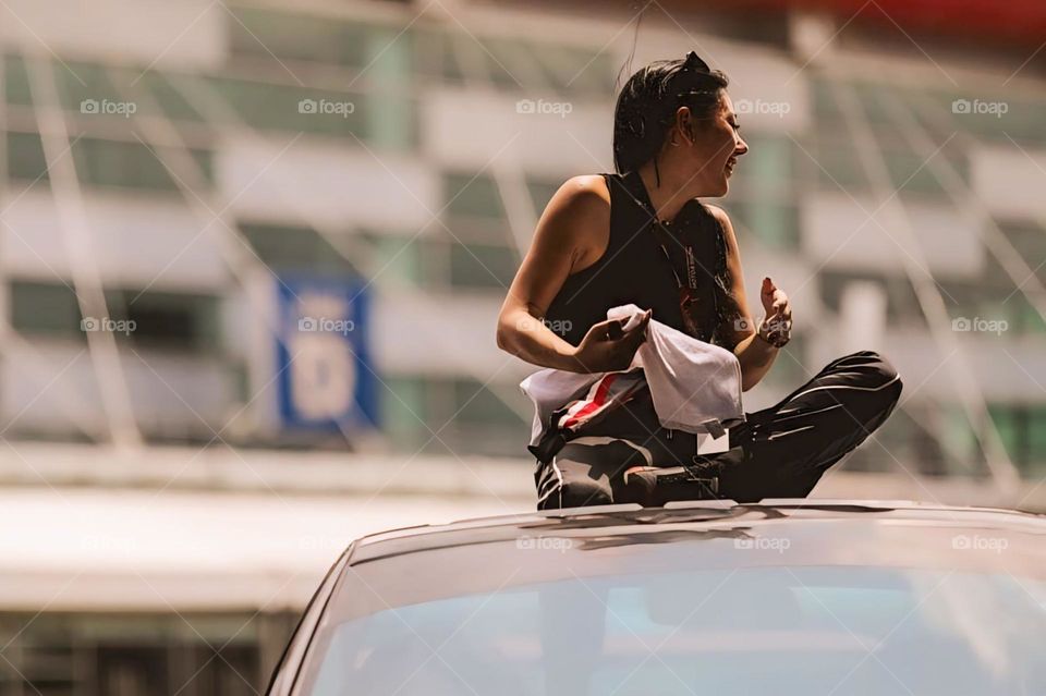 Portrait of a young female racer sitting on the roof of a car against the background of buildings and a clear sky