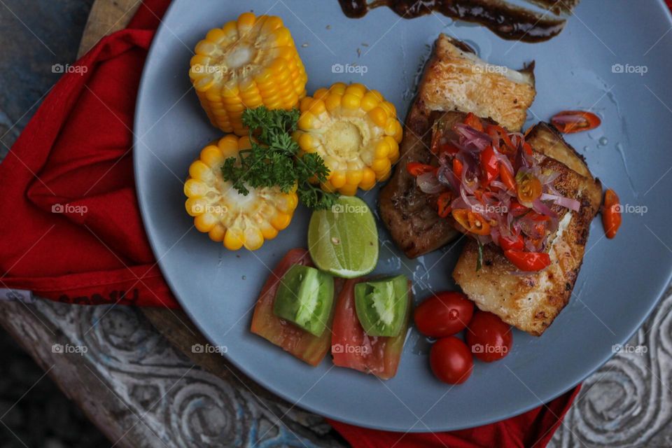 Tuna steak cooked with corn garnished with cherries, sliced tomatoes and lemon topped with sliced hot chili peppers on a baby blue melamine plate lined with red cloth and thick wood. Restaurant food.