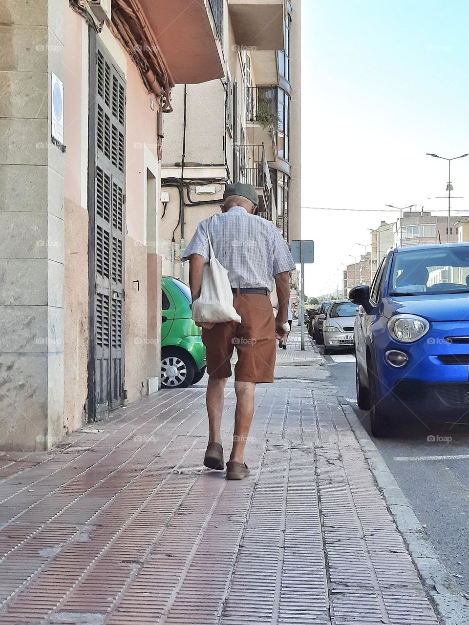 Elderly man walking down the street with his cloth bag and a treated wound on his right hand.