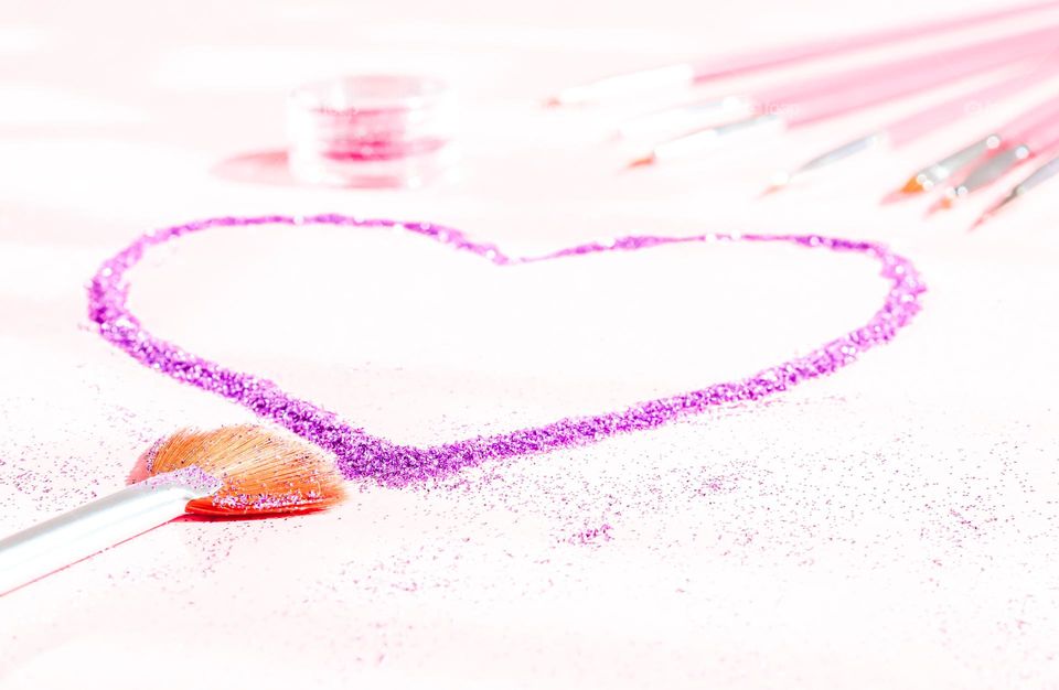 Lilac heart of glitter nail art with a set of brushes and a jar of glitter on a soft pink blurred background, side view. Concept cosmetics and beauty salons.