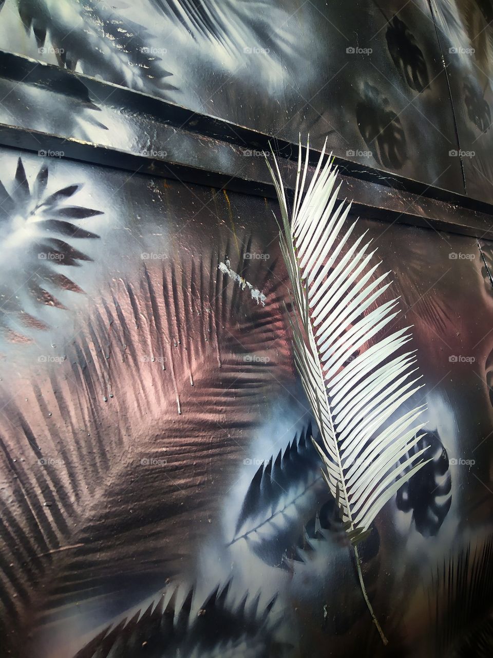 A palm leaf made of metal that is attached to a wall with graffiti in the form of leaf patterns