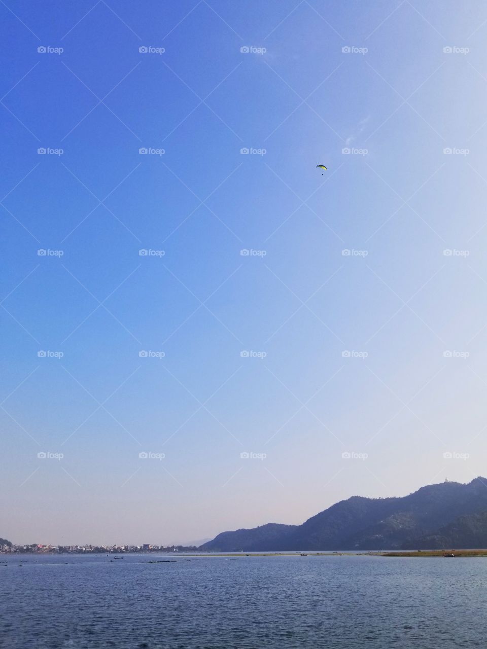 On the shores of Phewa Lake with a paraglider in the sky. The sky is a gorgeous blue which complements the blue in the foothills in the distance. It is almost a cloudless day.