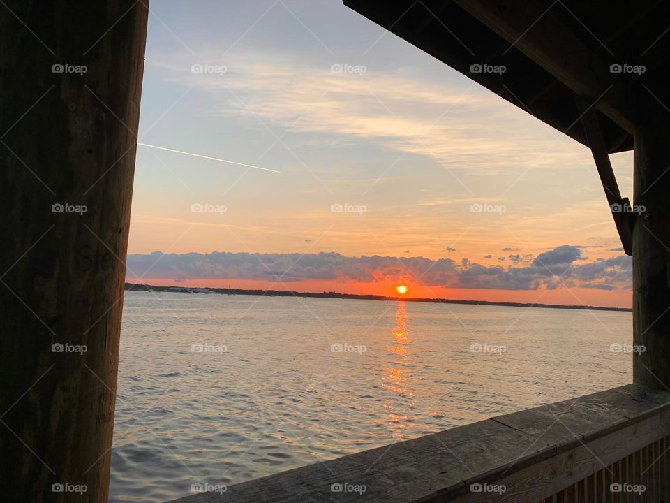 Sunset In The Horizon With Deep Orange Color Sighting From Gazebo On The Dock By The Island Reflecting On The Water.