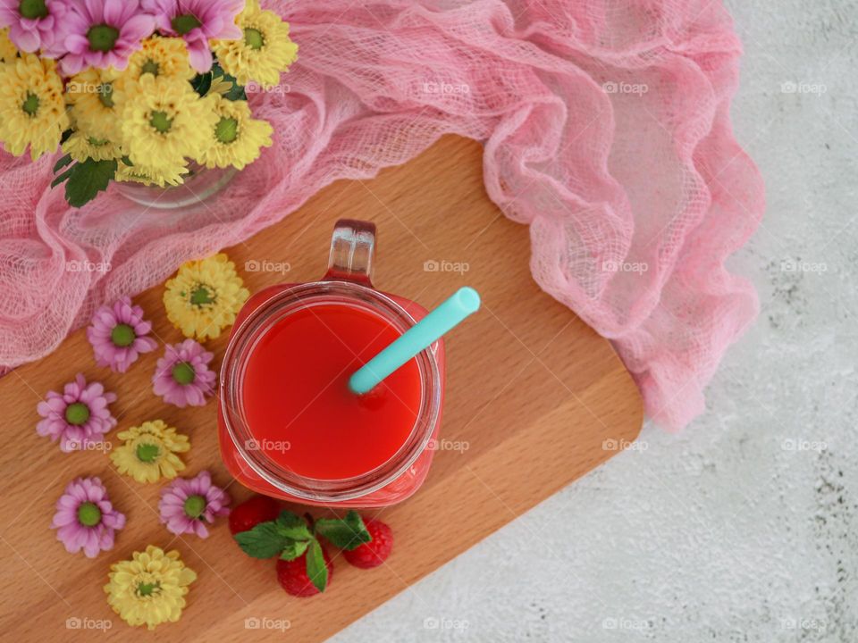 Natural strawberry drink in a glass with straws, a scattering of flowers with a bouquet in a mini vase stand on a wooden cutting board with a pink gauze napkin on a light concrete background, flat lay close-up. Summer drinks concept.