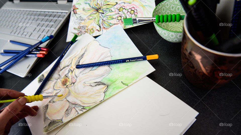 Closeup of hand holding Faber-Castell Art Grip Aquarelle Colored watercolor pencils sketching closeup spring flower with sketchbook on table with laptop and coffee conceptual work life balance, creativity, hobby artist lifestyle photography 