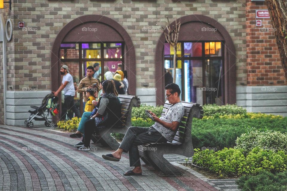 a man aged sits on a park bench looks into the phone. in the background are people and arched windows.