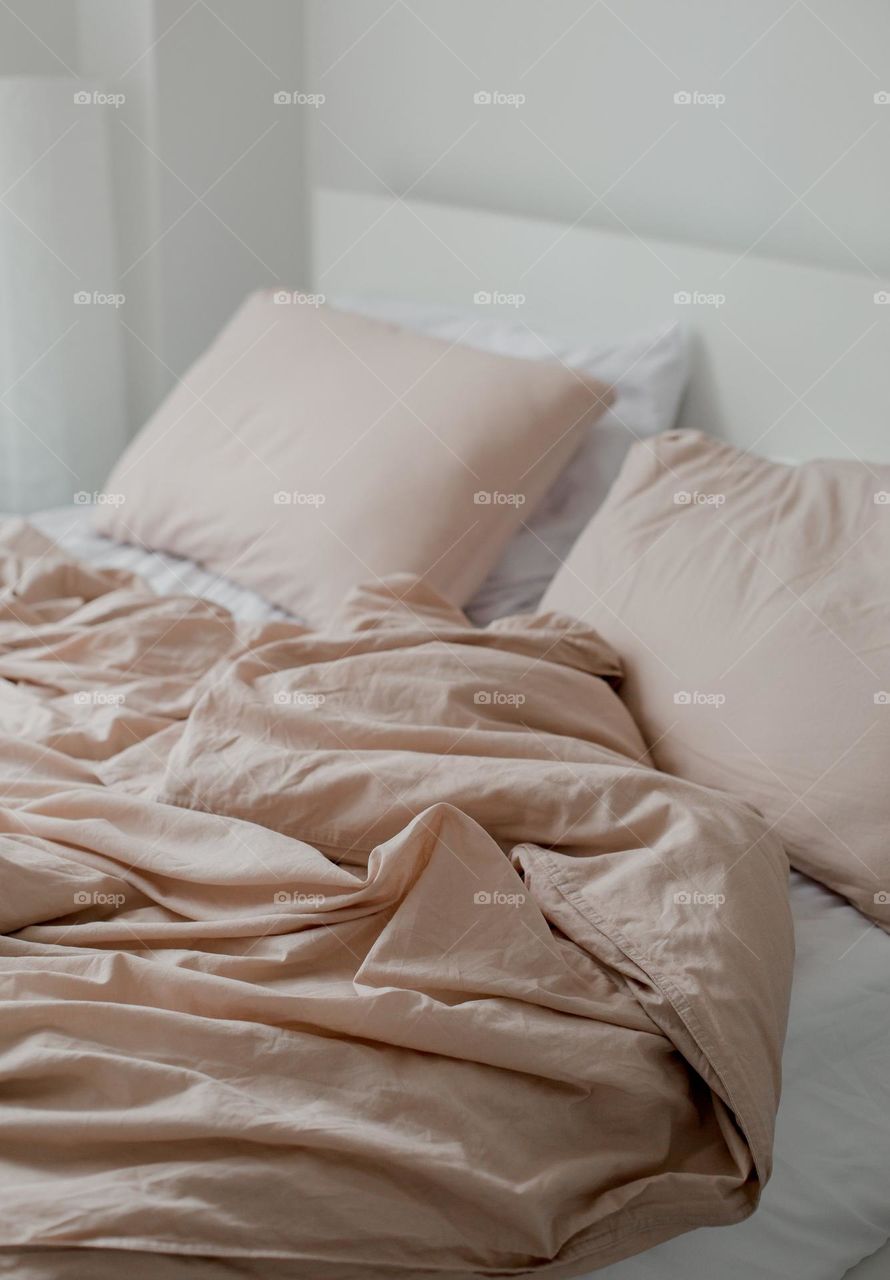 An unmade bed of dusty pink and white, with sunlight streaming gently  through a nearby window, hints at a restful night's sleep.