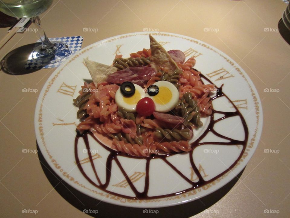 Cheshire cat shaped serving of rotini pasta, green and pink with hardboiled eggs and black olives for eyes with a small tomato nose and cornchip triangle ears.  served on a white plate with roman clock numerals and balsamic glaze garnish swirl.