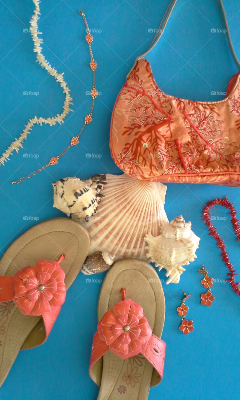 beach flatlay sandals seashells floral jewelry coral purse earrings ankle bracelet blue background vertical