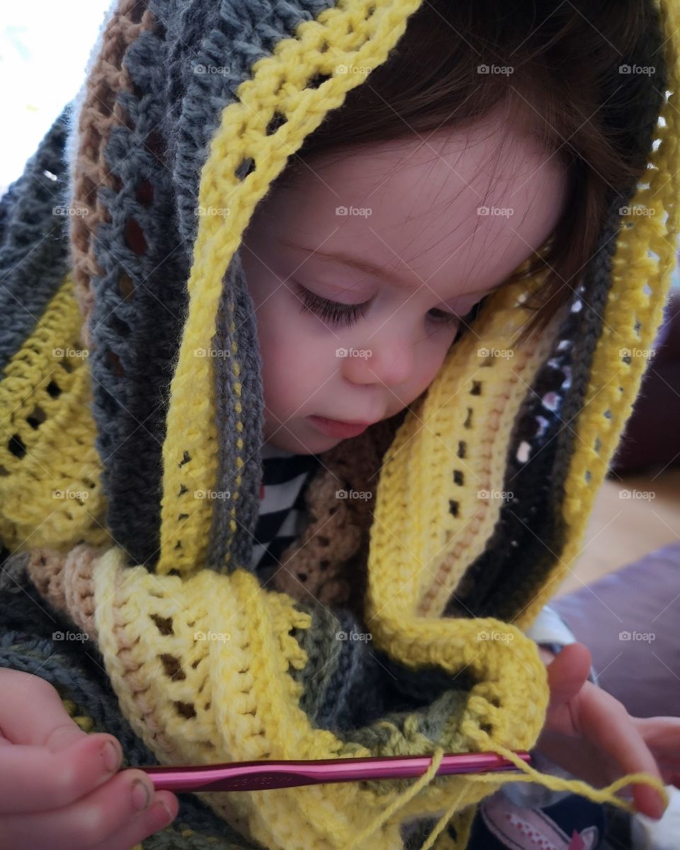 young toddler pretends to crochet while wearing a shawl on her head
