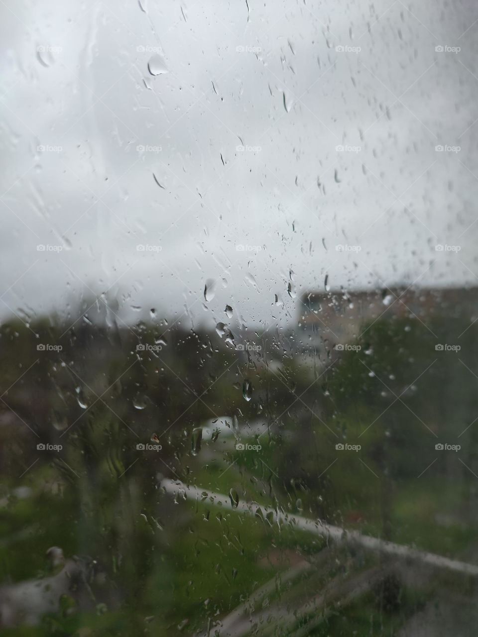 Raining day in the town, view from the window. Raindrops running on the window glass. Blurred skyline, trees, multi storey building at the background. Spring thunderstorm, gray heavy sky. View from the window, wet weather. Rain in the city, courtyard