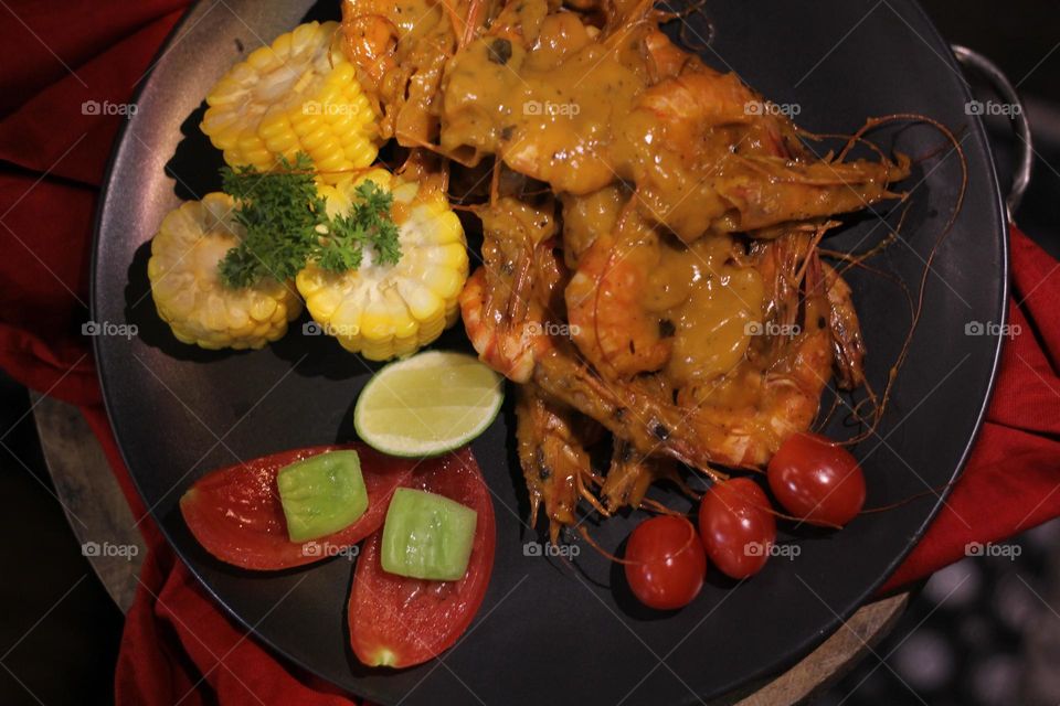 Asian food balado fried prawns with corn chunks, lemon slices and tomatoes served on black plate with red cloth bottom and wooden coaster. Asian style street seafood.