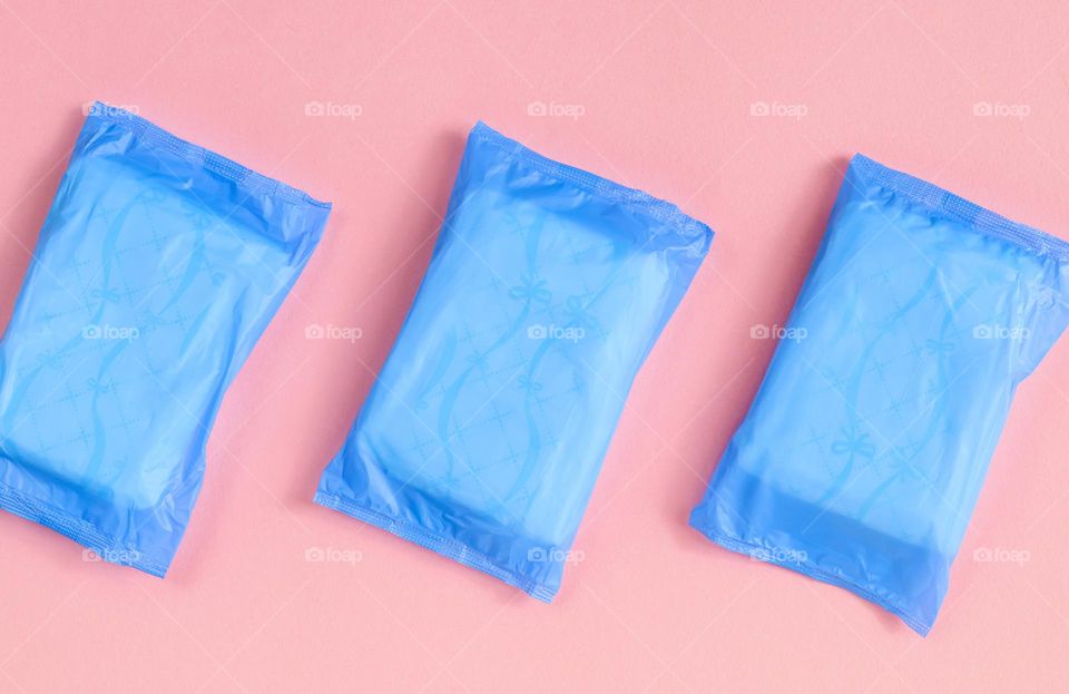 Three pieces of new women's pads in blue plastic packages lie rhythmically diagonally on a pink background, close-up side view. Health woman concept.