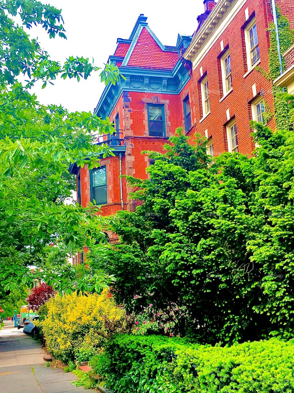 An old fashioned brownstone in Fort Greene, Bklyn with unusual decorative elements,a tower with a pointy steeple rooftop, delicate stucco ornaments painted blue in approx.the complimentary color to the red brick facade. 2019. Hypnotic Productions