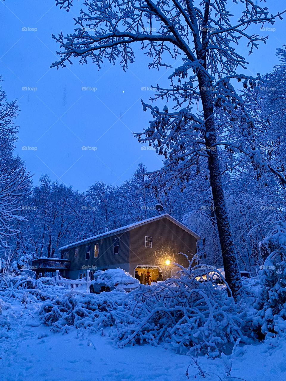Night during a snow storm in Northeast Pennsylvania USA; A house surrounded by snow covered woods and an open sky with the garage light on.