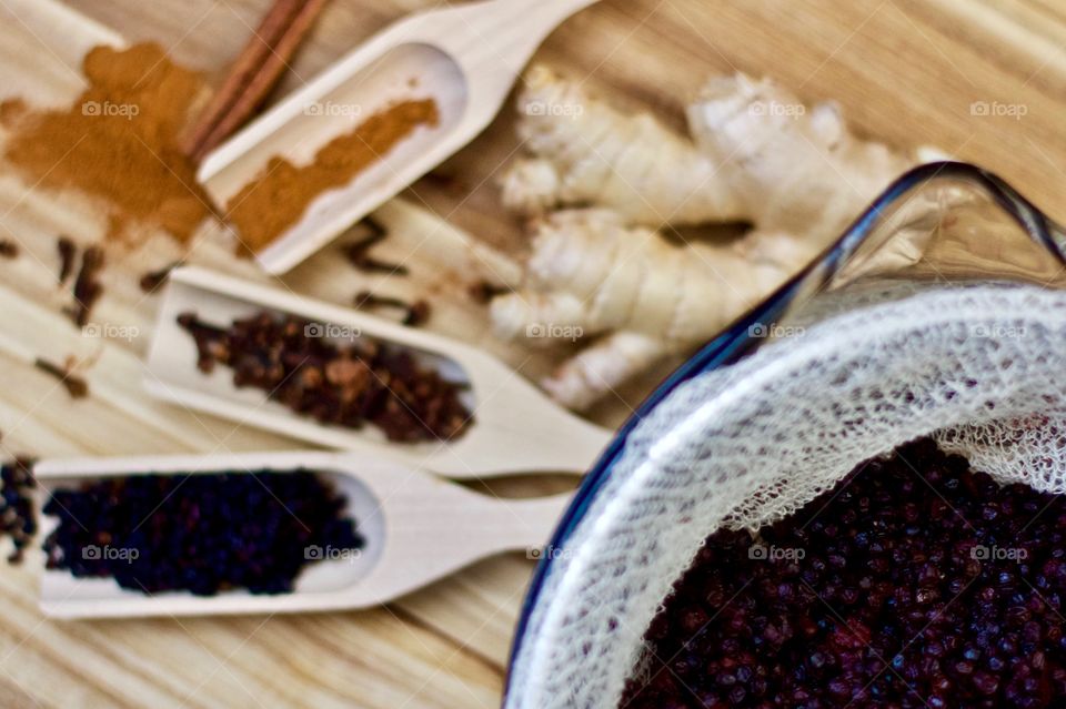 Overhead view of elderberries being strained for syrup in a colander lined with cheesecloth over a large glass measuring cup, blurred background including whole and ground spices in wooden scoops on a wooden surface