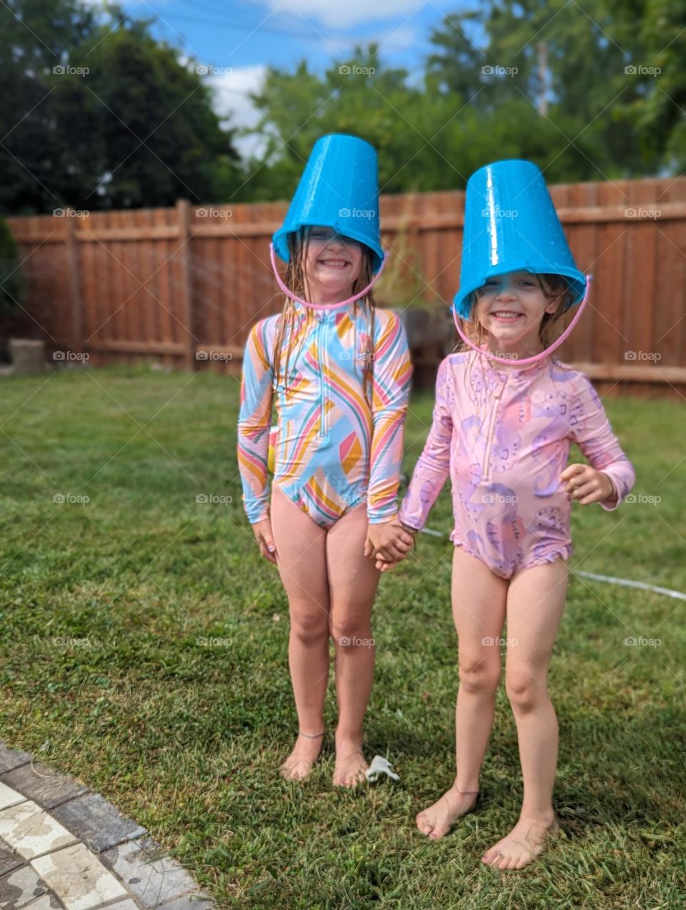 sisters playing outside in the sprinkler on a warm, sunny summer day, sand buckets on their heads and giant smiles, sisters holding hands, summer memories