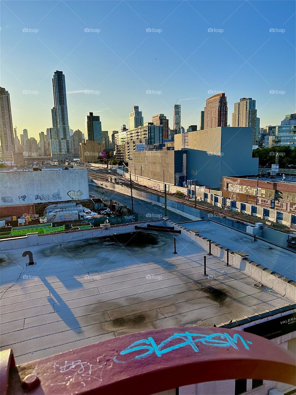 This is the view from the top of the red metal staircase of the “Pulaski Bridge” at “Newtown Creek” overlooking the “Long Island Railroad” tracks, “iFly”, an indoor flight simulator and “Manhattan” in the distance. 2024. Hypnotic Productions