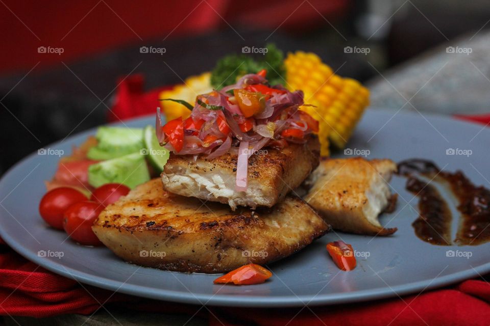 Tuna steak cooked with corn garnished with cherries, sliced tomatoes and lemon topped with sliced hot chili peppers on a baby blue melamine plate lined with red cloth and thick wood. Restaurant food.