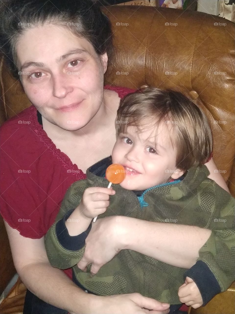 3-year-old son sitting with his mother licking an orange lolipop.