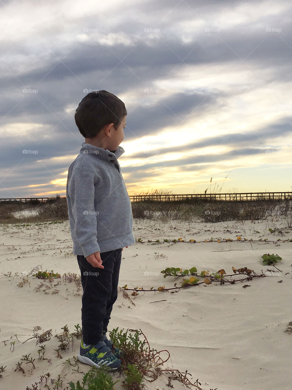 Young boy on sandy beach at sunset wearing sweater looking to horizon