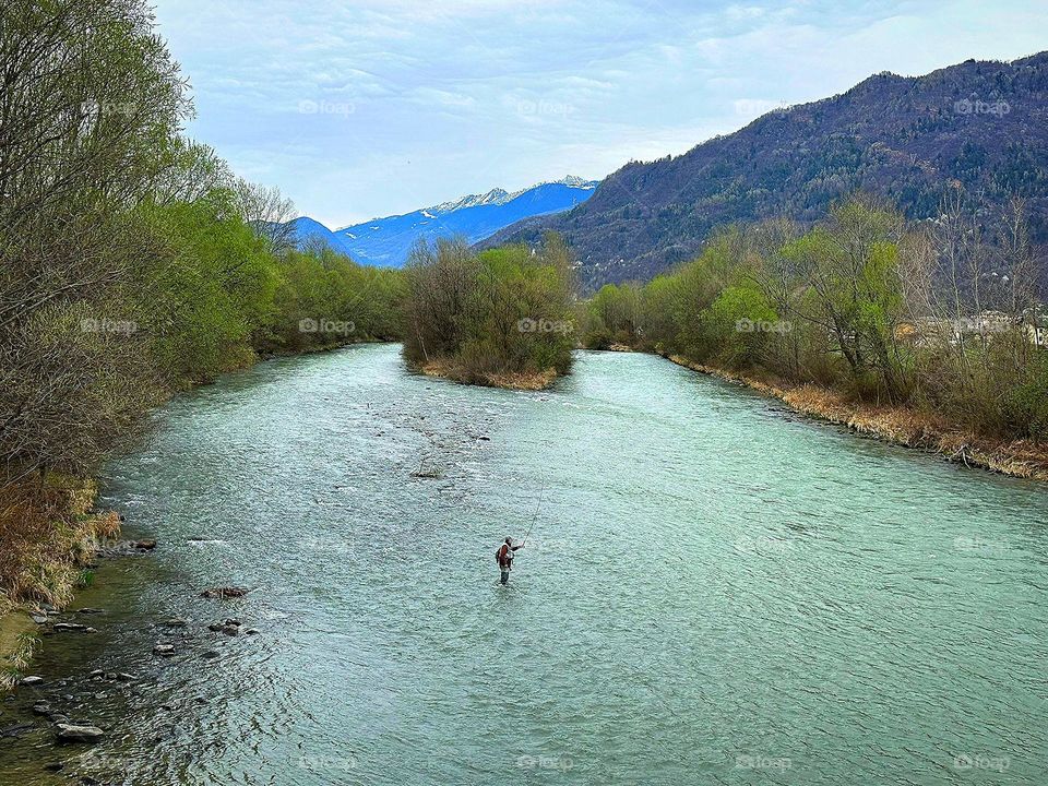 Spring river, which has become shallow.  There are green trees along the banks.  In the middle of the river stands a fisherman with a fishing rod and catches fish.  Mountain peaks are visible in the background.