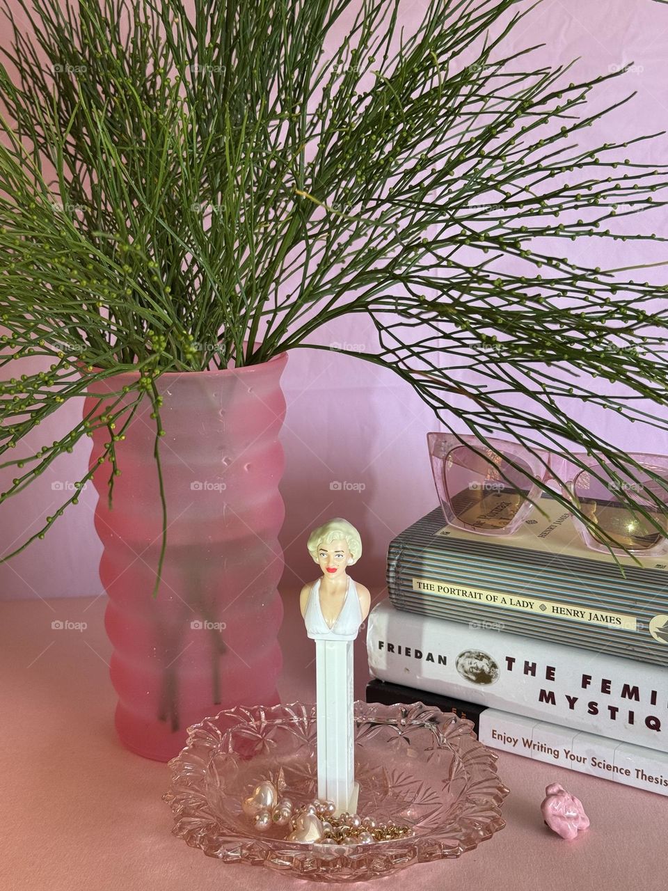 Marilyn Monroe Pez dispenser in a pink glass trinket dish with pearl earrings with a wavy vase holding whisk fern (Psilotum nudum) fronds with a stack of books, a pair of pink plastic sunglasses and a porcelain chewed bubblegum pin