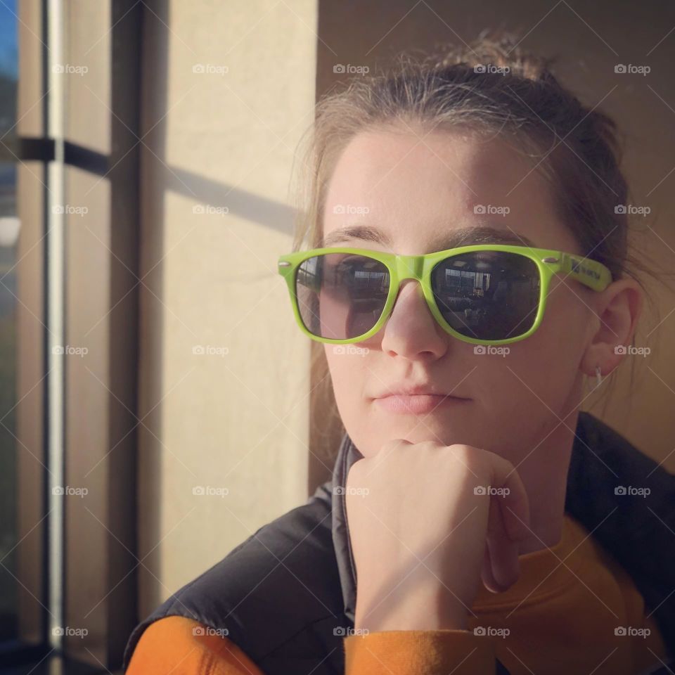 A vibrant young girl in sunglasses sits near a window, bright light illuminating her face 