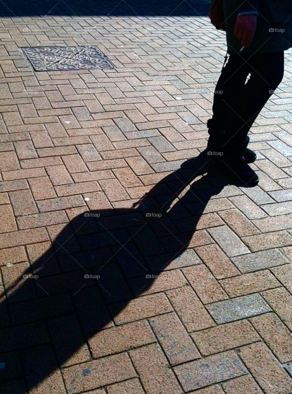Bright sunshine causing solid shadow from a lady standing on patterned pavement. 