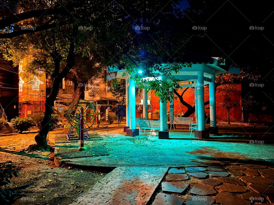 An empty park, with four benches under a canopy. Beside the canopy is colourful exercise equipment. It is nighttime, and a turquoise light shines on the park.