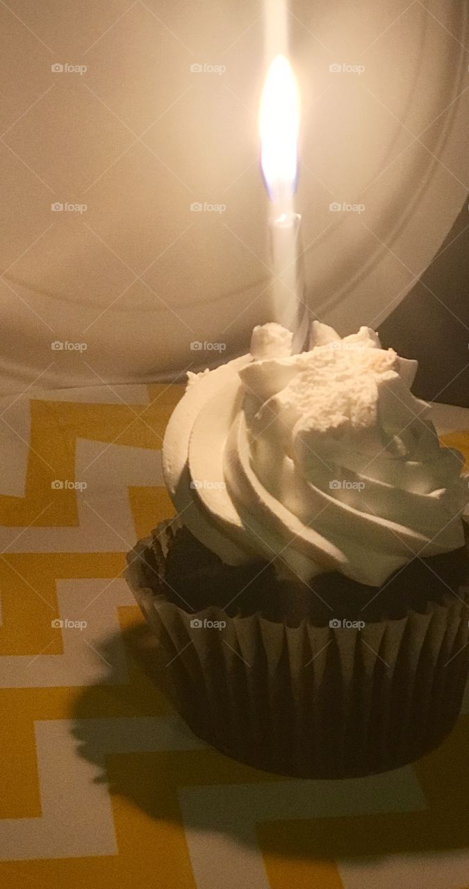 Vanilla frosting on a yummy chocolate cupcake with a bright and shiny glowing candle on a yellow checkered paper Birthday plate