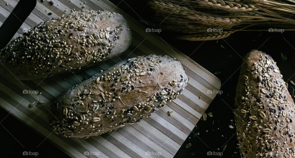 2 seeded rolls on a wooden board with a sheaf of wheat dimly lit from one side