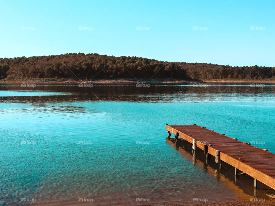 Looking across a lake from the end of a small pier to a small tree covered mountain. 