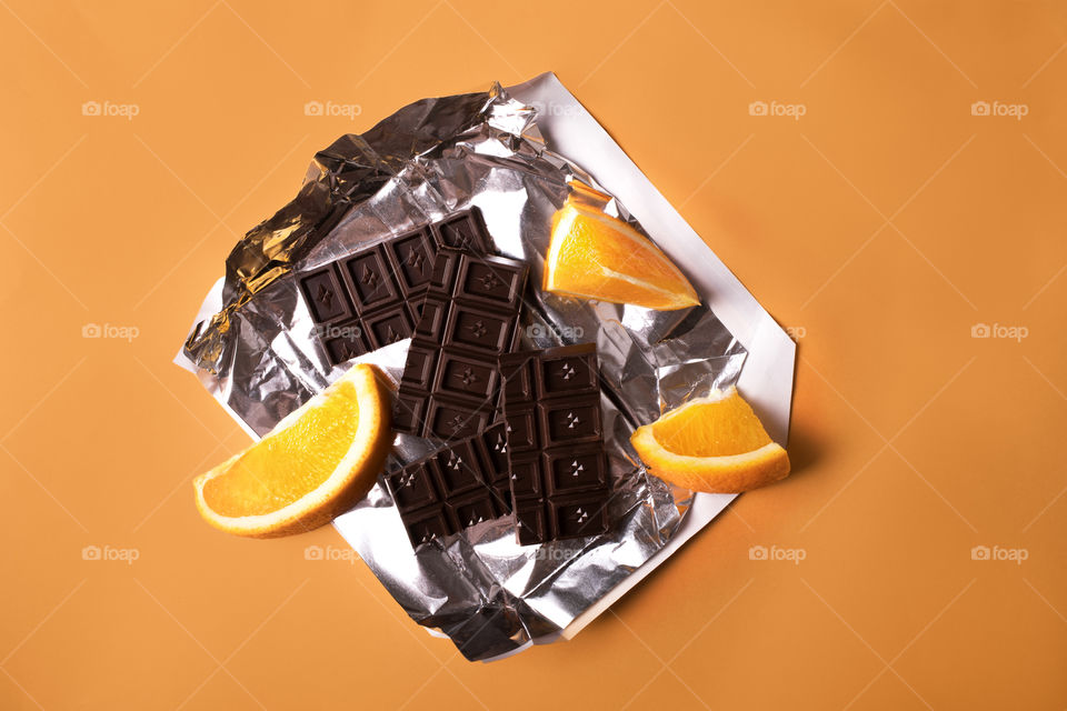 Chocolate bar broken in pieces with orange segments on foil insert an paper package in yellow background