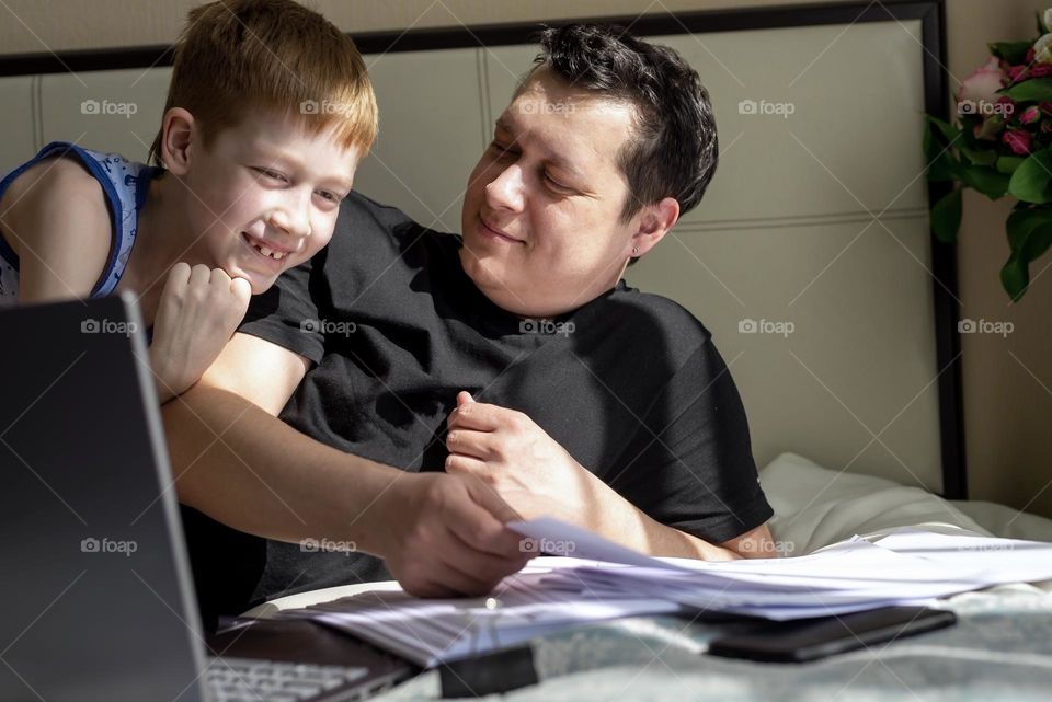 Father and son spend fun time together, father works at home and son sit next to the bed with a laptop, close-up lifestyle portrait