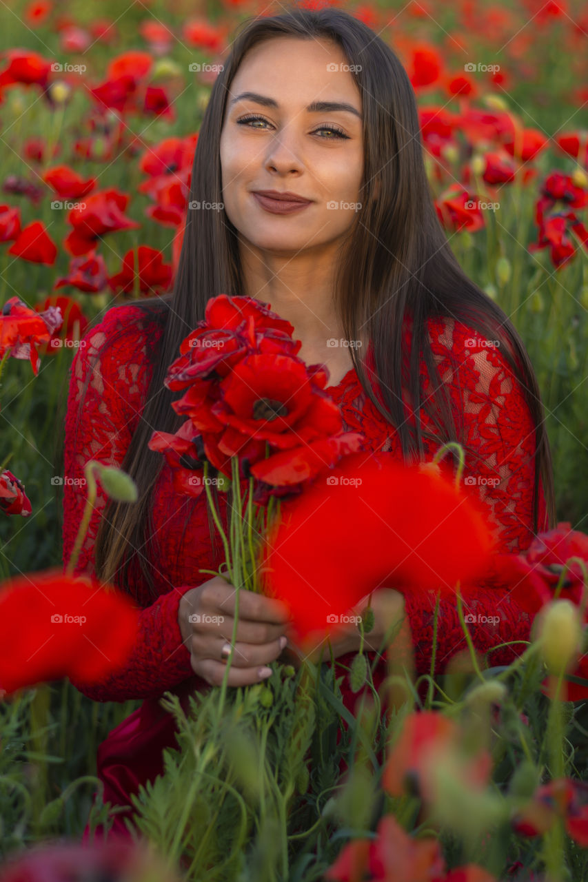 Close-up front view of a beautiful woman with  perfect clean face with a bouquet of poppies sitting in a poppy field and looking directly at the camera in nature