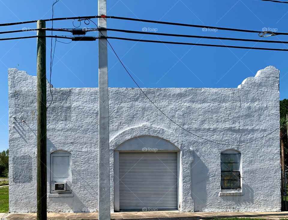 Old white architectural building with signs of aging elements under a blue sky in the afternoon with turned edges arched garage door and windows on both sides.