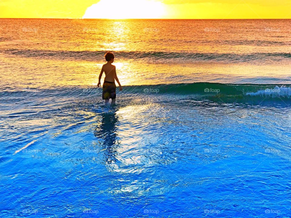Young boy wading through blue waves under a spectacular golden sunset.