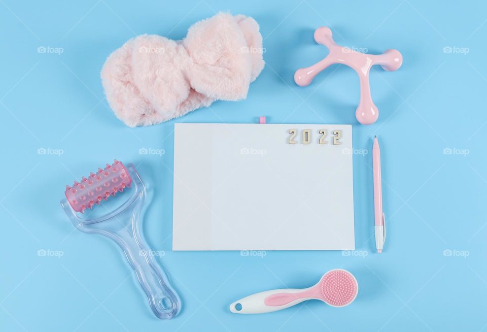 Set for female massage: brushes, massager, pink fluffy headband with ears, empty notepad, pen and wooden numbers 2022 on blue yon, flat lay close-up.